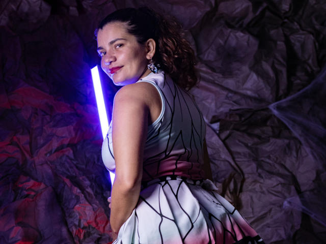 BulmaHot with curly brown hair, wearing a white dress with black and pink geometric patterns, holding a purple light, smiling, looking over her shoulder, showing her ass.