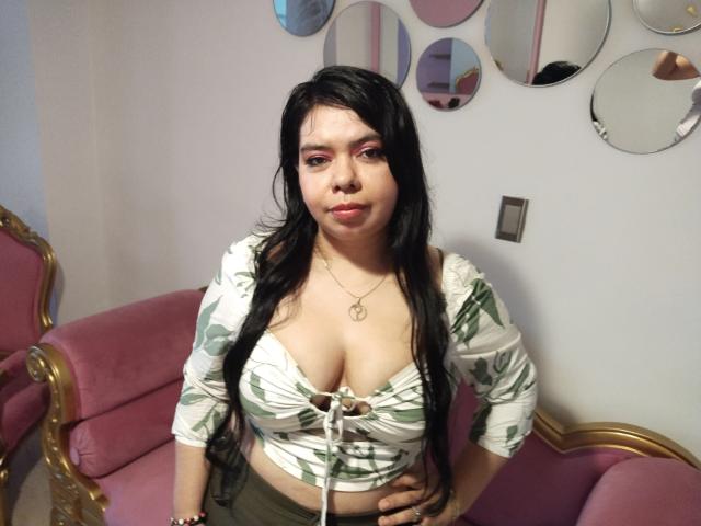 KarlaSexyHott with long black hair, pink eye makeup, white crop top with green leaf pattern, lace-up front, showing her big tits, sitting on pink velvet chair.