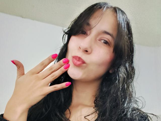 AnneStone with black wavy hair, pink lipstick, and pink nail polish, kissing her hand, showing off her tits and ass with a cute, sexy expression.