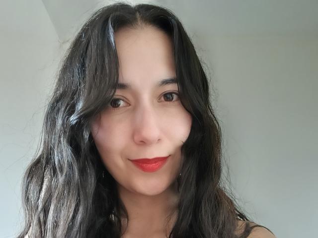 AnneStone with dark, wavy hair, red lipstick, and light skin, smiling slightly, looking directly at the camera. Her tits are not visible, just her face and upper chest.