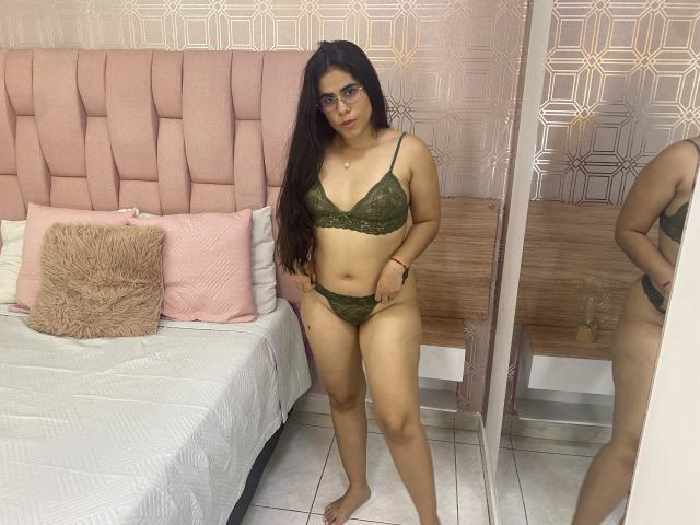 AlicyaTorres stands in a bedroom wearing green lace lingerie, showcasing her medium-sized tits and curvy ass. She has long dark hair, glasses, and a neutral expression.
