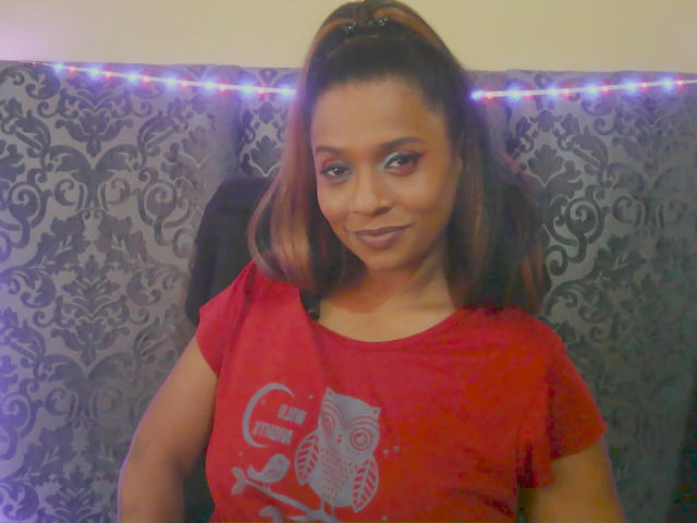 MiZzCreator with brown ponytail, red shirt, and owl graphic, smiling, showing tits, brown eyes, and subtle makeup, standing in front of a dark patterned curtain with blue lights.