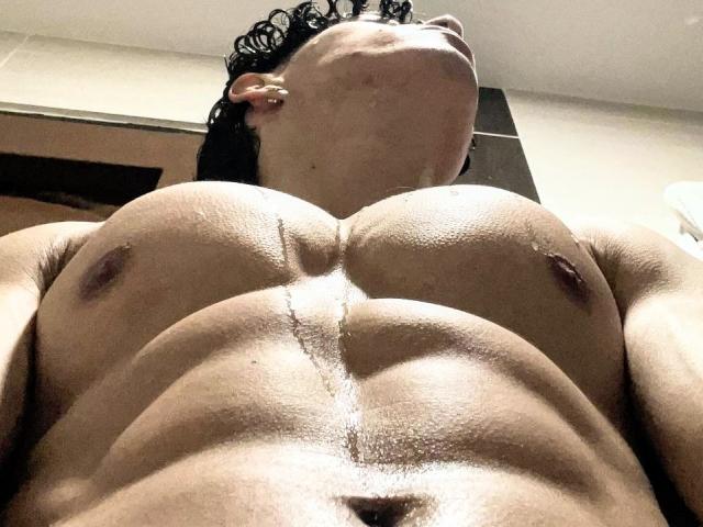 NoahJhoness, muscular, shirtless, low-angle, wet skin, visible nipples, defined abs, chest, short curly hair, nose ring, head tilted back, slight shadow on right, skin glistening.