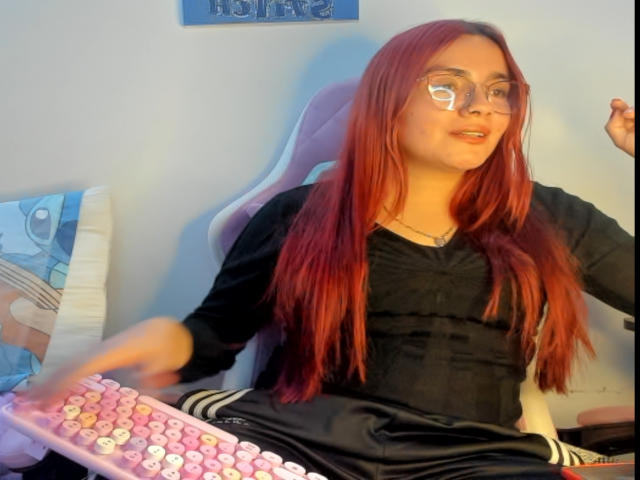 EimmyHoskX with red hair, glasses, black shirt, pink keyboard, sitting on a pink chair, right hand raised, left hand on keyboard, subtle smile, relaxed, casual, home setting.