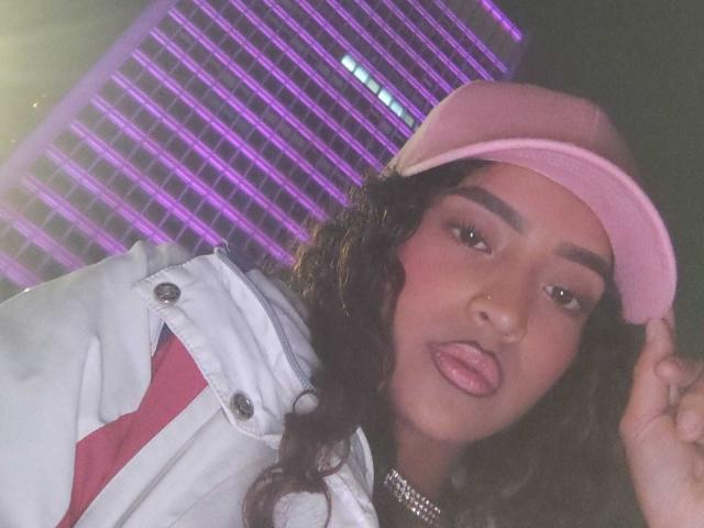 ValeryMonroe with long curly black hair, pink baseball cap, white jacket with red stripes, and silver choker, sticking out pink tongue, looking directly at camera, in dim light.