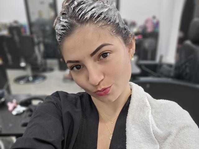 KatherineToro with wet, silver-tipped hair, black top, white towel, gold necklace, and pink lipstick, looking slightly to the side, in a hair salon.