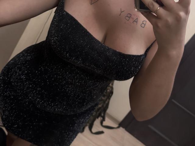 Kametia's sparkly black dress shows her big boobs and a tattoo on her chest. She's taking a mirror selfie with her phone, her hand partially in frame.