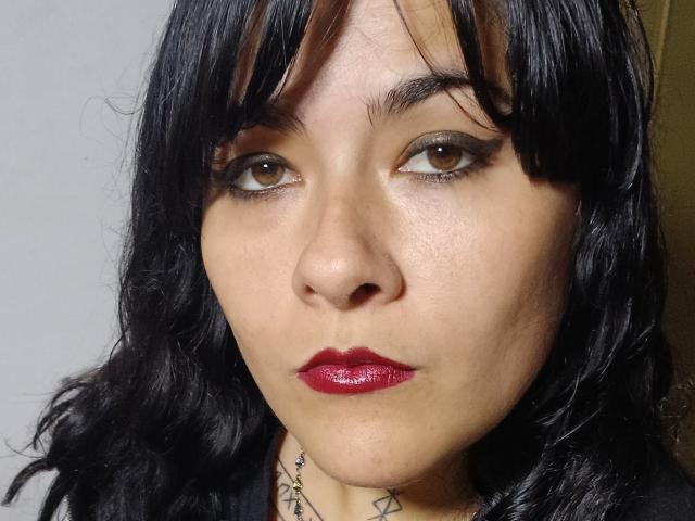 FreeMystic69 tiene el cabello oscuro y ondulado, ojos marrones con delineador de ojos ahumado y labios rojos y brillantes. Lleva una blusa negra y un collar geométrico..