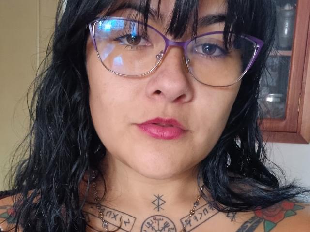 FreeMystic69 tiene el pelo negro y ondulado, gafas moradas, lápiz labial rosa y tatuajes en el pecho. Tiene un pequeño aro en la nariz. Está mirando directamente a la cámara con una expresión neutra..