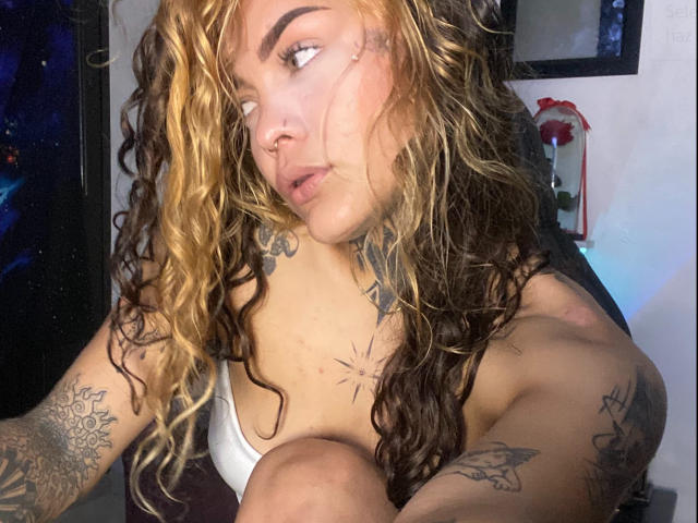 KyraX with curly blonde and brown hair, nose ring, and tattoos, looking to the side, showing small tits in a white bra, sitting with knees up.