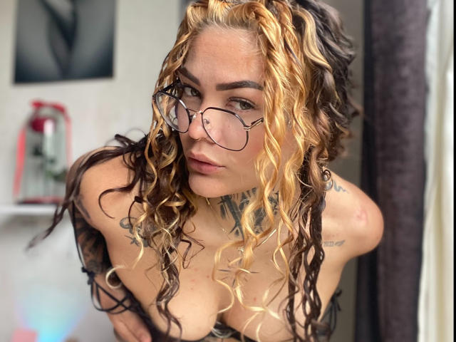 KyraX with curly blonde and brown hair, wearing glasses, leaning forward, showing her tits, tattoos on her chest and arms, looking into the camera.