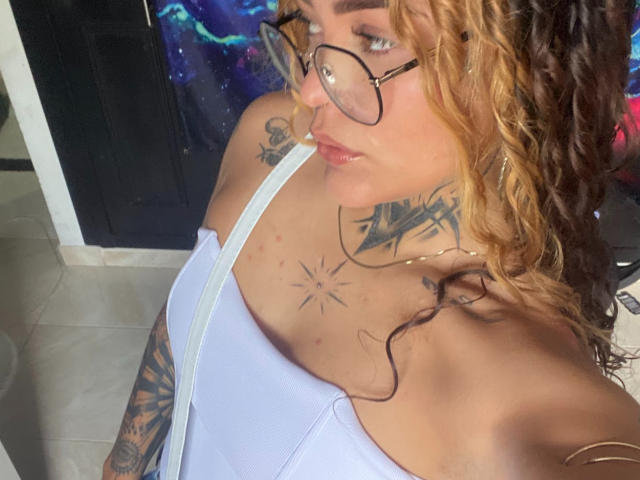 KyraX with curly blonde and brown hair, glasses, white tank top, tattoos on chest and arm, looking up, pale skin, small nose, pouty lips, visible bra strap.