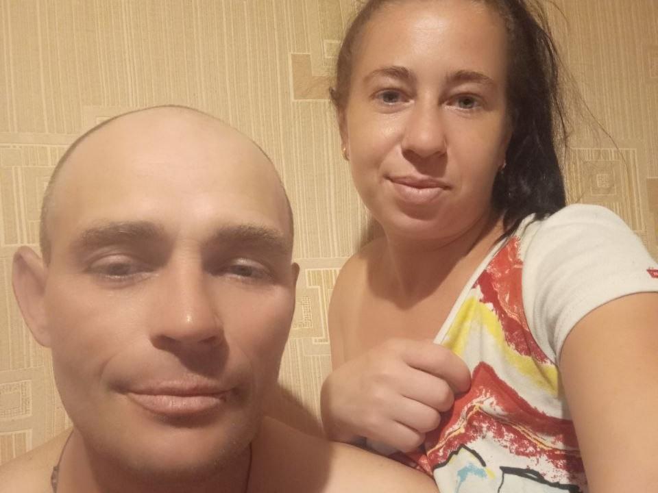 TonyAlina, avec la tête rasée, et TonyAlina, avec les cheveux bruns, souriant, tenant une chemise colorée, montrant ses seins, debout ensemble, épaules nues, peau claire, éclairage chaleureux, selfie décontracté..
