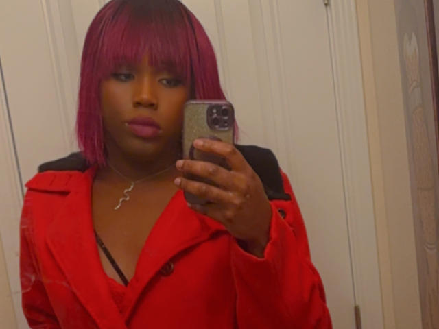 Jinkx4You with purple bob, red jacket, black straps, holding phone, necklace, pink lipstick, looking at mirror, medium brown skin, holding phone in right hand.