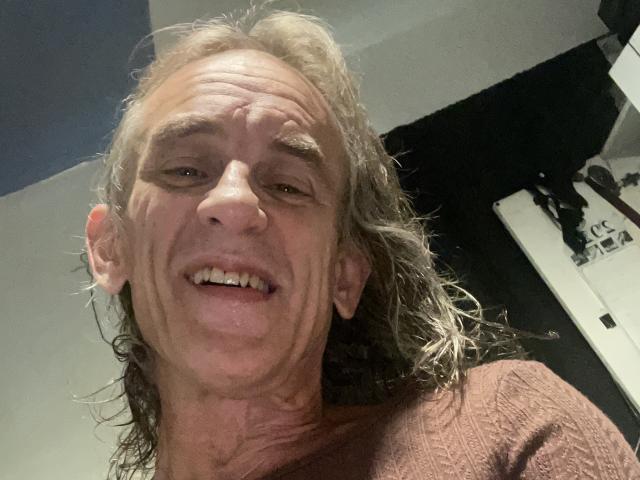 Middle-aged Supermodel69 with long, wavy, grayish-brown hair, wrinkled face, and slight smile, wearing a brown, textured shirt. No visible genitals or boobs.