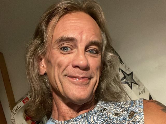 Supermodel69 has gray, shoulder-length hair, blue eyes, and a slight smile with visible teeth. Wearing a blue, patterned shirt. Tattoo on right shoulder. Older, handsome dude.