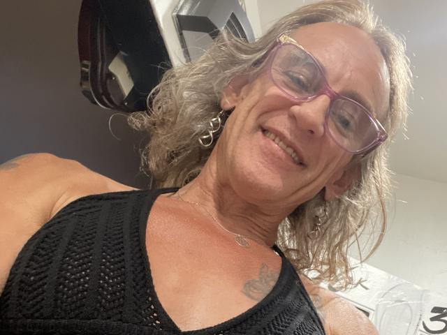 Supermodel69 with gray curly hair, purple glasses, black patterned top, smiling, showing tattoos on shoulder and chest, wearing hoop earrings, and a necklace.