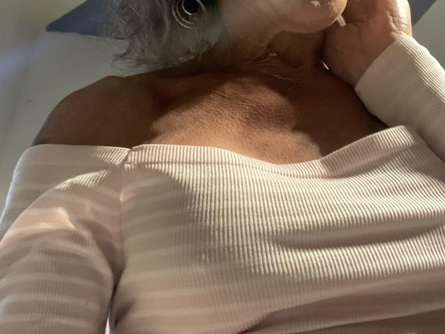 Supermodel69's tits are visible under a white, off-shoulder, ribbed top. Her hand is near her mouth, with gray curly hair and a gold necklace.