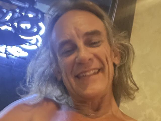 Supermodel69 with gray, wavy hair, smiling, showing wrinkles and sagging skin, topless, revealing small, sagging tits, in a slightly blurry, indoor photograph.