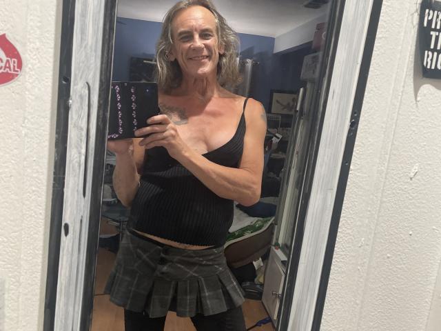 Supermodel69 with long gray hair, smiling, wearing a black ribbed tank top, showing small tits, and a gray plaid skirt, holding a phone in a mirror selfie.