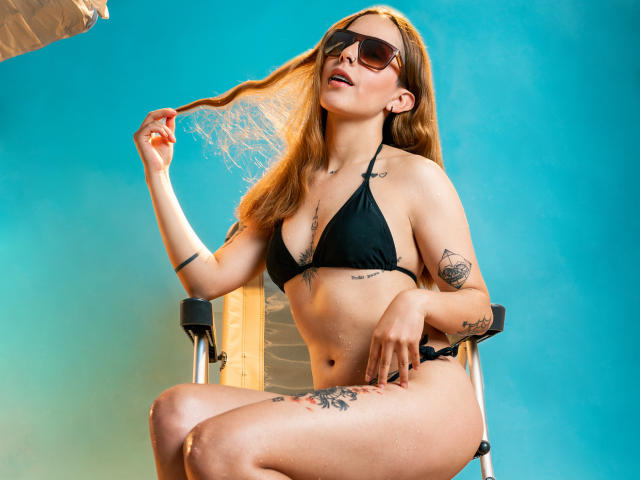 VictoriaOrtega with long blonde hair, black bikini, sunglasses, tattoos, holding hair, sitting on chair, blue background, looking up, hand on hip, showing her tits and ass.