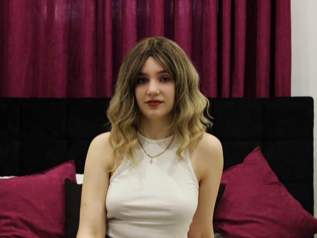 MilenaAudley with wavy blonde hair, wearing a white sleeveless top, sitting on a bed with purple pillows, and a black headboard, looking at the camera with a slight smile.