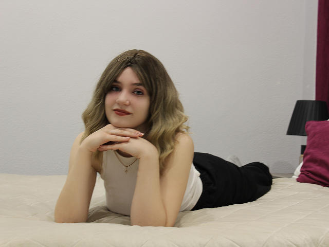 MilenaAudley, lying on a bed, white tank top, black pants, blonde hair, red lipstick, hands under chin, smiling, pale skin, small tits, relaxed, bedroom, white wall, black lamp.