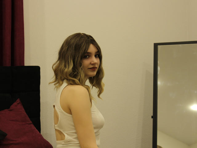 MilenaAudley with wavy blonde hair, white tank top showing sideboob, looking over her shoulder, standing near a mirror, bedroom setting, dark red pillow, black headboard.