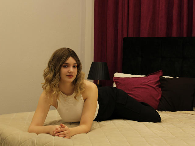 MilenaAudley, blonde woman with wavy hair, lying on beige bed, wearing white sleeveless top and black pants, looking at camera, hands under chin, red and purple pillows in background.