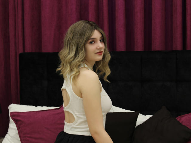 MilenaAudley with blonde wavy hair, wearing a white sleeveless top with cutouts, black skirt, sitting on a bed with purple and black pillows, holding a black laptop.