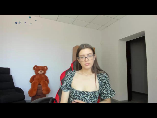 DoraLi with glasses, long brown hair, wearing a floral dress with a deep cleavage, sitting in a chair, brown teddy bear on the left, white room. Boobs visible.