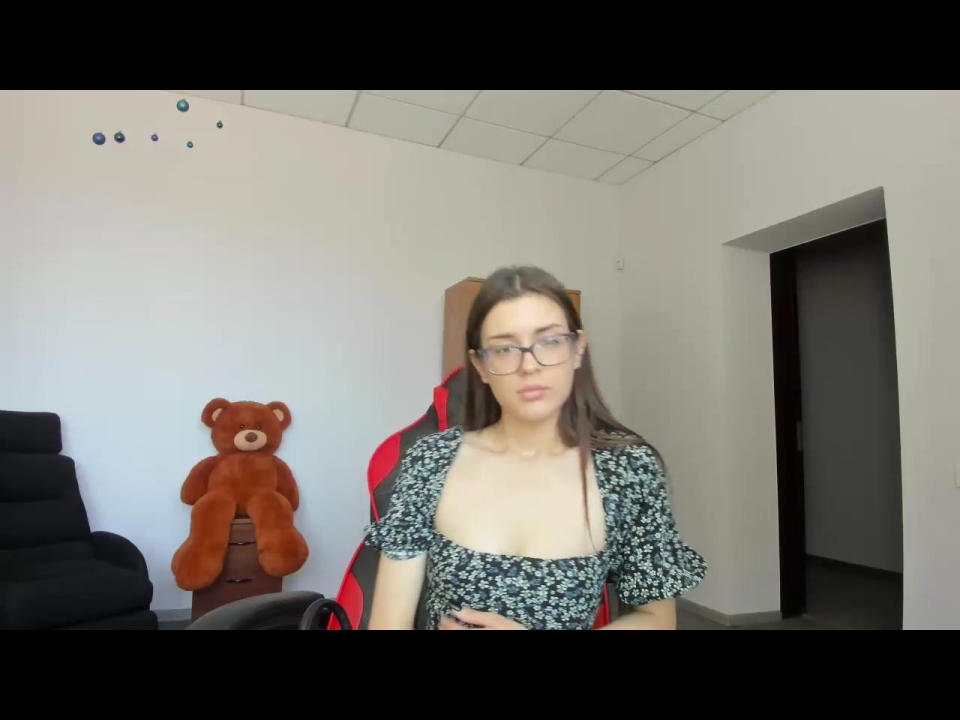 DoraLi with glasses, long brown hair, wearing a floral dress with a deep cleavage, sitting in a chair, brown teddy bear on the left, white room. Boobs visible.