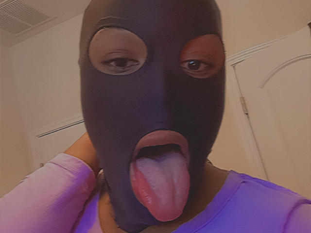 Jinkx4You wearing a black mask with eyes and open mouth, tongue out, in a pink shirt. Their mask covers their face, eyes looking forward.