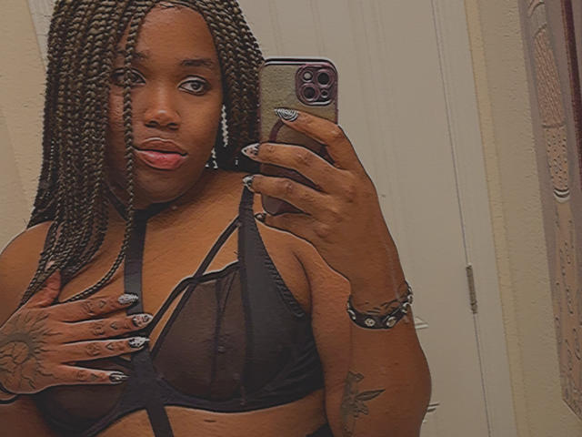 Jinkx4You with braided hair, dark skin, black sheer bra, tongue out, holding phone, hand covering tits, tattoos on hand and wrist, black nail polish, studded bracelet.