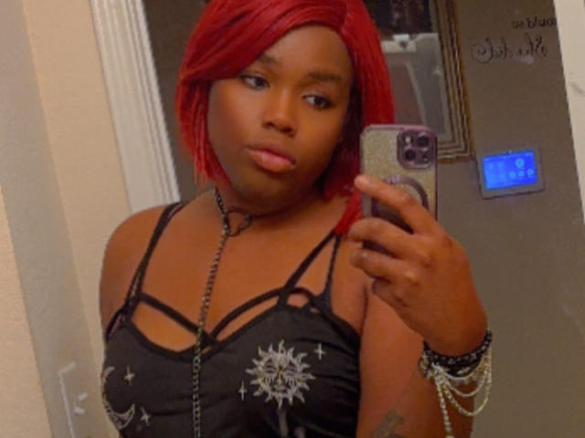 Jinkx4You with red hair, dark skin, pouty lips, wearing a black strappy top, holding a phone, looking in a mirror, with bracelets on her right wrist.