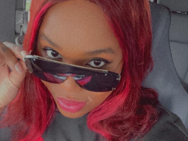 Jinkx4You with red hair, holding black sunglasses in front of her face, showing dark eyes, pink lipstick, and a slight smile, sitting in a car.