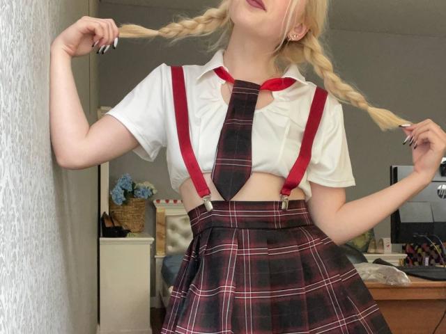 LaPulia met blonde vlechten, rode braces en een geruite rok, die haar borsten laat zien onder een wit shirt en een rode das, en die haar vlechten vasthoudt. Slaapkamer setting..