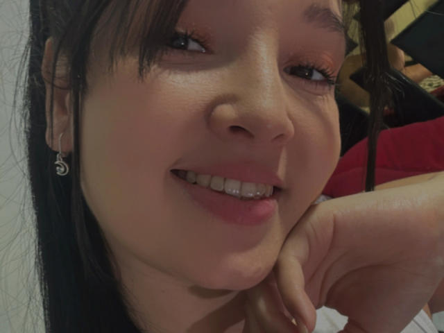 SarahGood has a cute smile with pink lips, brown eyes, and black hair, resting her chin on her hand. She's wearing small silver earrings and has a slightly shiny face.