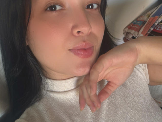 SarahGood's face with black hair, light skin, pink lips, and brown eyes, wearing a white turtleneck, hand near chin, slightly pouty expression. Boobs not visible, ass not visible.