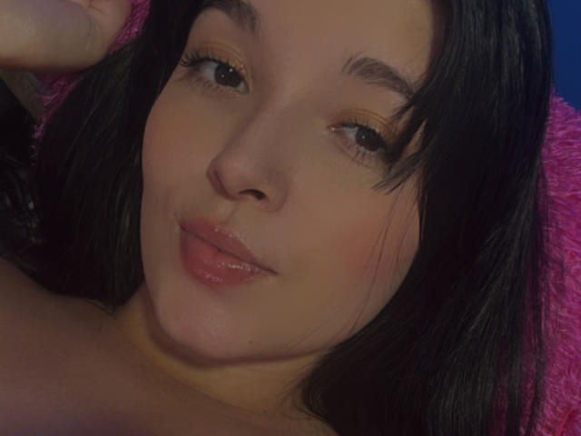 SarahGood has glossy pink lips, dark brown eyes with long eyelashes, and black hair. Her hand rests near her head. She's lying on a pink, fuzzy surface.