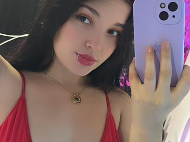 SarahGood with dark hair, pink lips, and a gold necklace, holding a purple phone, wearing a red dress, taking a selfie, showing her tits, tongue out.