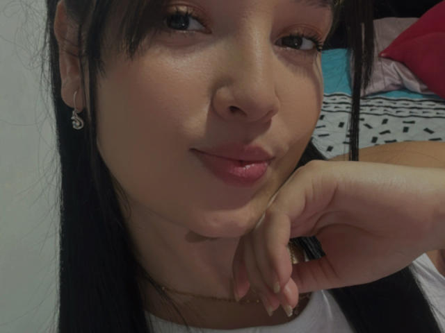 SarahGood's face with dark hair, brown eyes, and pink lips, resting her chin on her hand, wearing a white top and gold necklace, with small earrings.