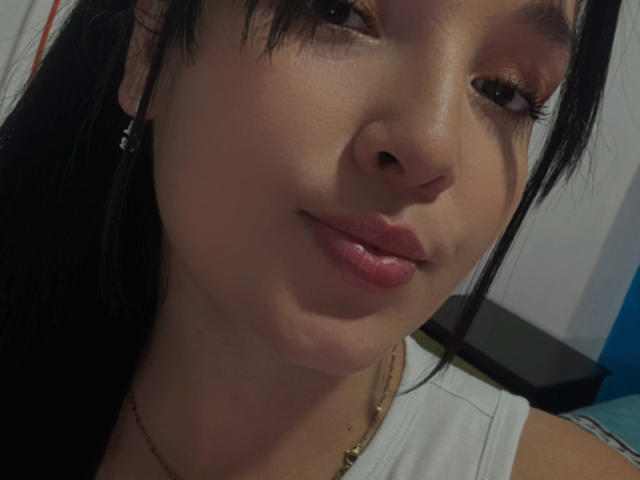 SarahGood has a cute face with brown eyes, black hair, and pink lips. She's wearing a white top and a gold necklace, with a slight smile on her face.
