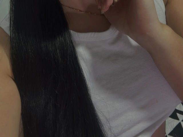 SarahGood with long black hair, wearing a white tank top, touching her chin with her right hand. Her chest is visible but not the focus.