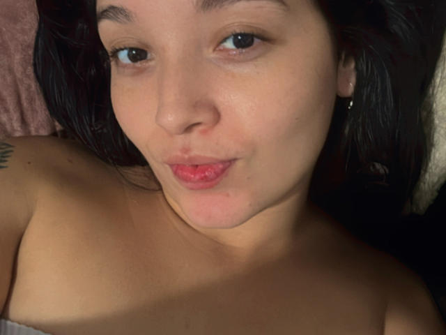 SarahGood's face, pouty lips slightly puckered, dark hair, light skin, small tattoo on left shoulder, blue eyes, wearing earrings, topless, black background, subtle light source from front.