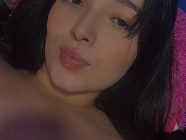 SarahGood's face with dark hair, glossy pink lips, and light brown skin, looking slightly up with a subtle smile, her bare shoulders visible.