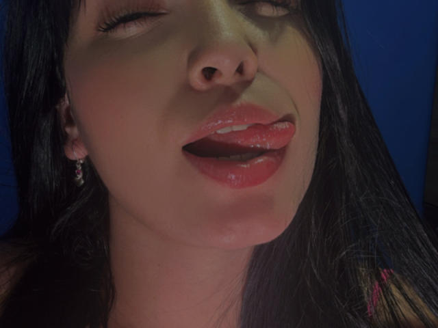 SarahGood's face, eyes closed, tongue out, glossy pink lips, long black hair, small hoop earrings, slightly open mouth, blue background, soft lighting, slight blush on her cheeks.