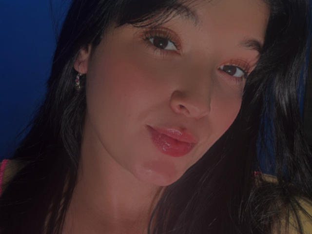 SarahGood's face with shiny pink lips, dark brown eyes, and black hair. She's wearing small earrings, looking slightly to the right with a subtle smile. Her skin is light.