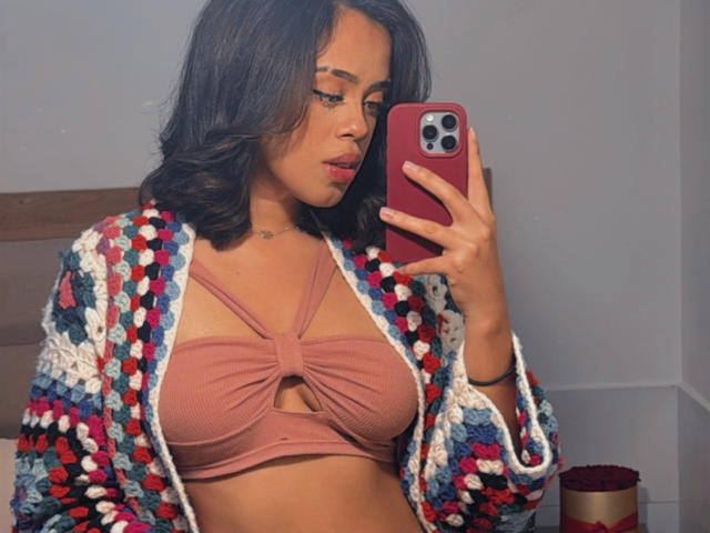 Lia Hilton, avec une poitrine de taille moyenne, portant un soutien-gorge rose avec une découpe en forme de clé de voûte, et un cardigan coloré en crochet, prenant un selfie devant un miroir avec un téléphone rouge..