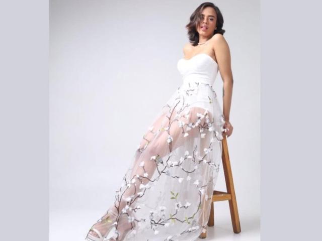 LiaHilton with white strapless dress, sheer floral overlay, sitting on wooden stool, smiling, wavy brown hair, white background, dress flows to the floor, soft pink and white flowers.